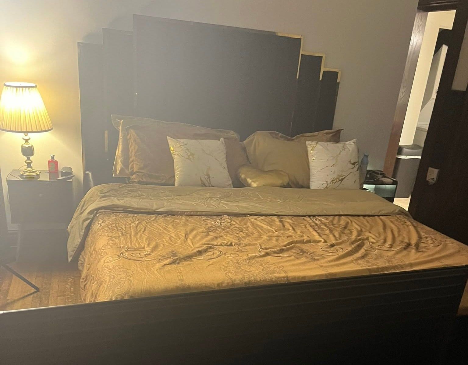 A bedroom after cleaning, with a gold bedspread neatly laid, pillows arranged and a lamp lit on the nightstand.